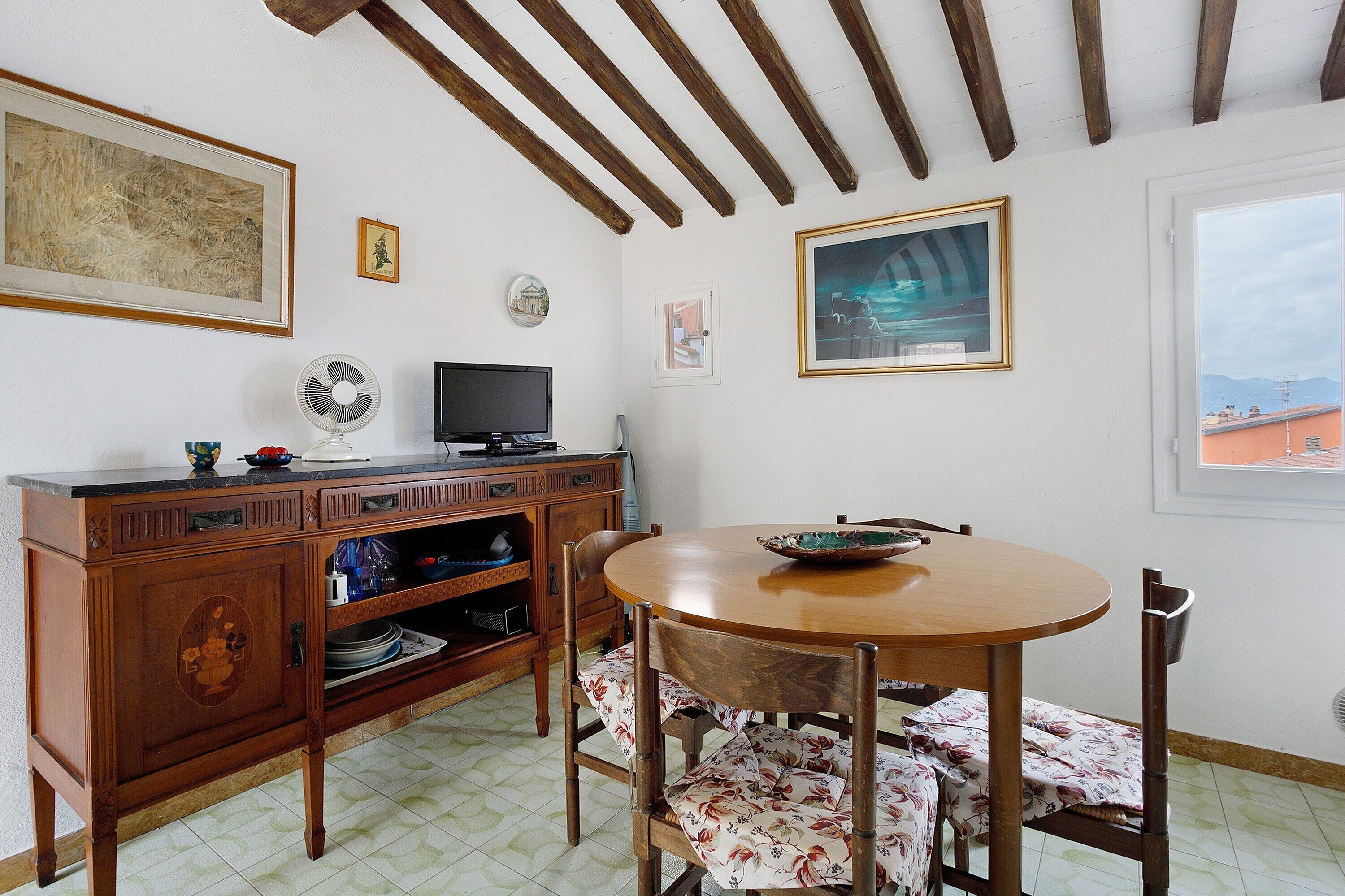 A dining area with a round wooden table, chairs with floral upholstery, a wooden cabinet with shelves, a television, and framed artwork on the walls.