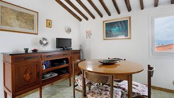A dining area with a round wooden table, chairs with floral upholstery, a wooden cabinet with shelves, a television, and framed artwork on the walls.