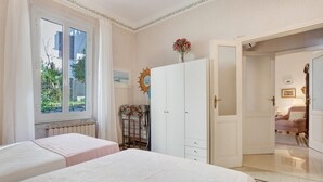Ein Schlafzimmer mit zwei Betten, einem Kleiderschrank, einem Heizkörper und einem Fenster mit Blick auf Grünflächen.