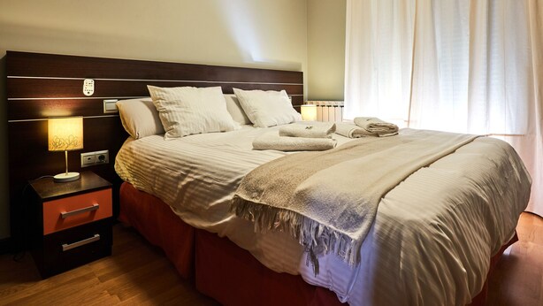 A neatly made bed with pillows and a blanket, a bedside table with a lamp, and a nightstand with drawers. - AV Soterra a 2B in Vila (Ávila)