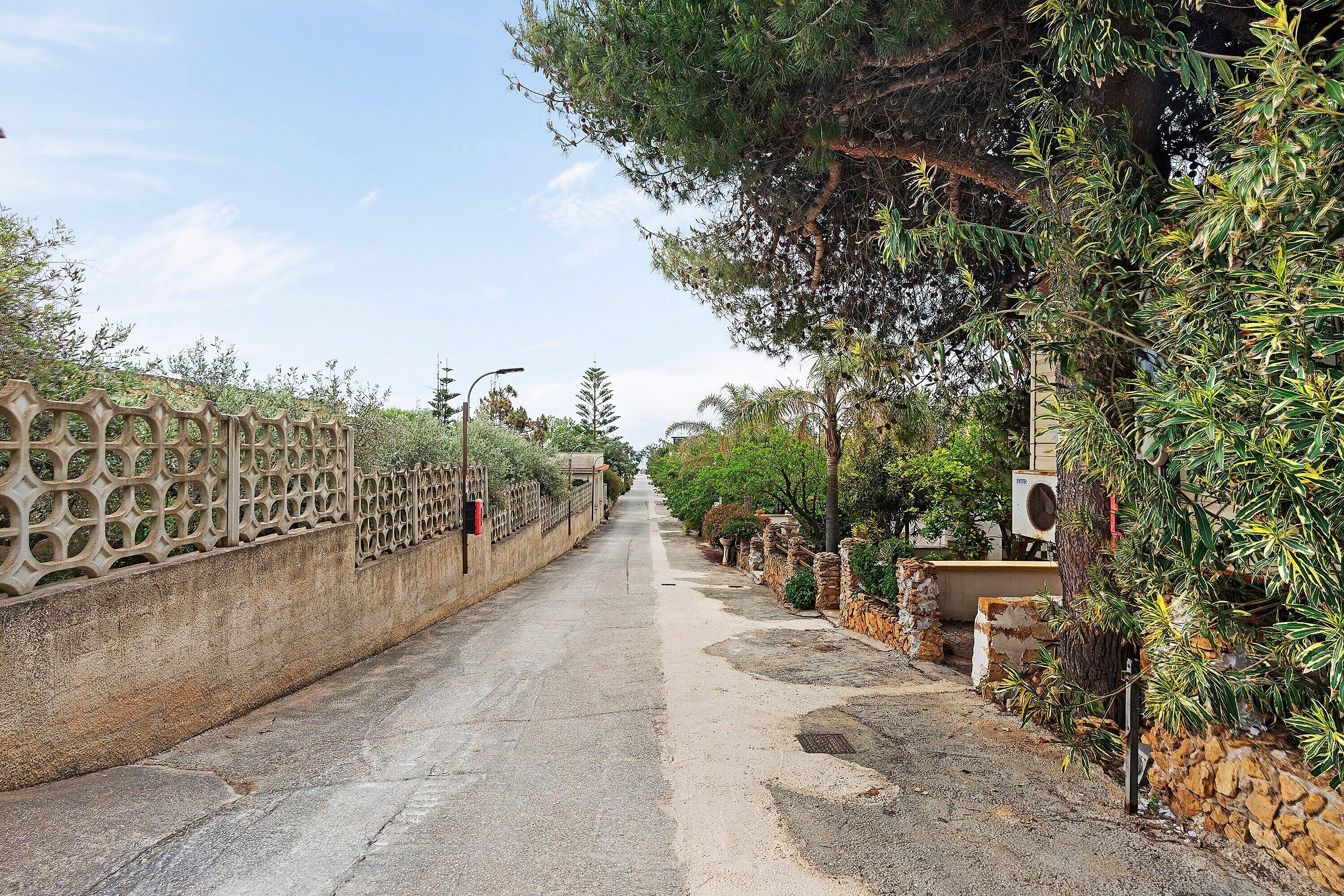 A paved pathway flanked by a decorative fence and stone walls, leading to a building with a tree and an air conditioning unit.