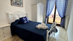A neatly made bed with a blue bedspread and pillows, a white bedside table with drawers, a white wardrobe, and a window with blue curtains. - From Pupa SeaView Apartment (Vico Equense)