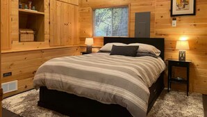 A wooden cabin-style bedroom with a bed, two bedside tables with lamps, built-in shelving, and a window with blinds. - Serene 1-bedroom cabin in charming Ashford perfect for getaway (Ashford)