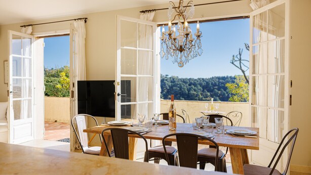 Een eetruimte met een tafel voor vier personen, een kroonluchter en uitzicht op een bebost landschap door open deuren.
