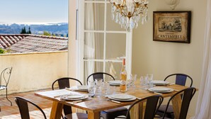 Een eettafel voor zes personen, gedekt met borden, glazen en een fles wijn, met uitzicht op een pannendak en bergen.