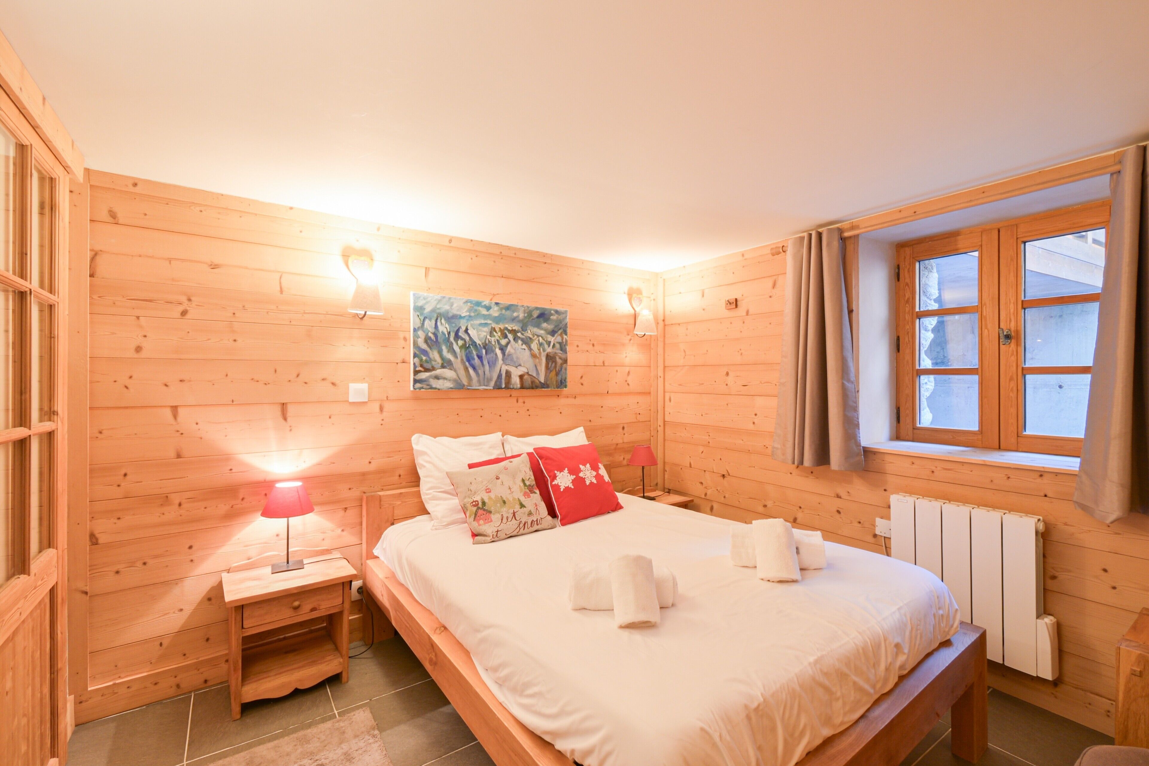 Een kamer met houten lambrisering, een bed, nachtkastje, lamp en een schilderij aan de muur.