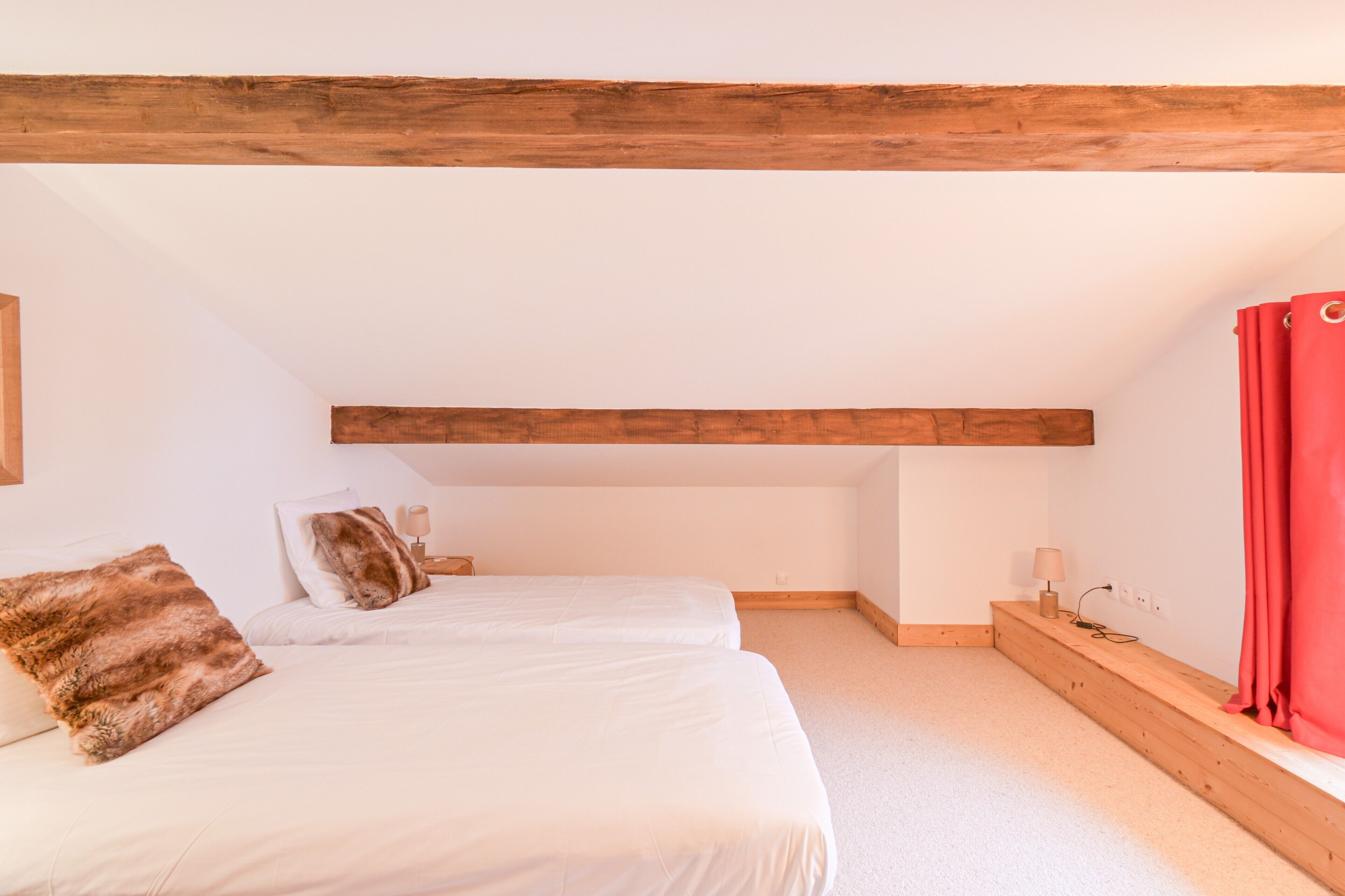 Een slaapkamer met twee bedden, houten balken, een rood gordijn en een klein nachtkastje met een lamp.