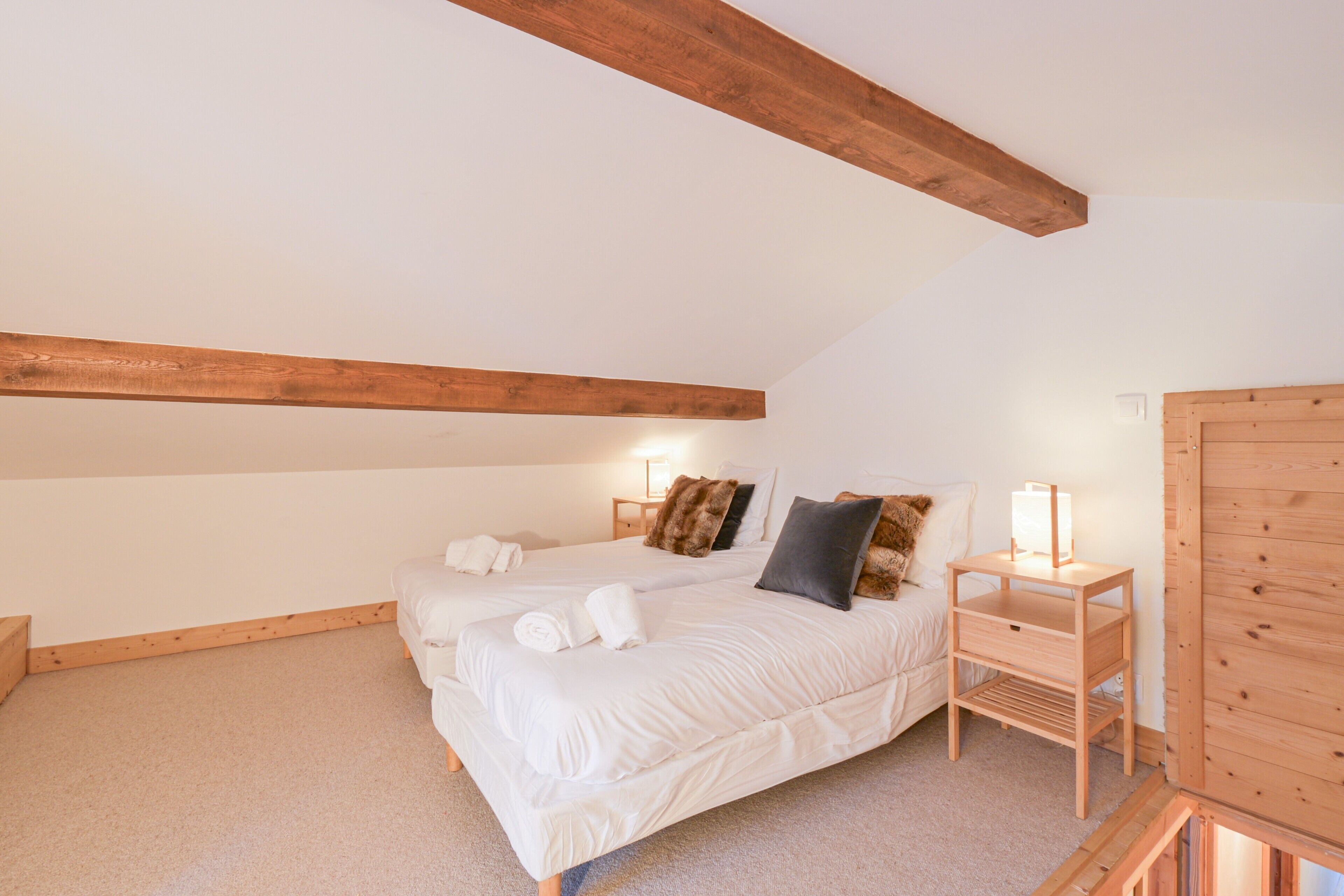 A bedroom with two beds, wooden beams, a nightstand with a lamp, and a wooden wardrobe.