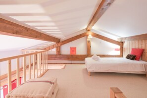 Une chambre avec un plafond en pente, des poutres en bois, un lit avec une literie blanche, un oreiller rouge et un petit tabouret en bois.