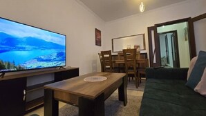 A living room with a flat-screen TV, a wooden coffee table, a green sofa, and a dining area with wooden chairs and a table.