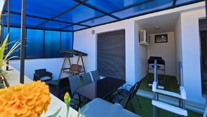 A covered outdoor seating area with a table and chairs, a swing set, and a small garden.