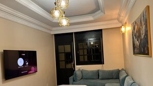 Un salon avec un téléviseur à écran plat fixé au mur, une table basse blanche, un canapé d’angle gris et un tapis à motifs.