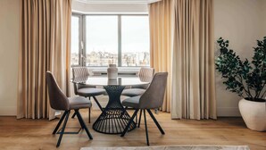 A dining area with a round table and four chairs, a potted plant, and a view of a cityscape through large windows. - The Eccleston: Elegant 2BR Flat in Prime Belgravia (London)
