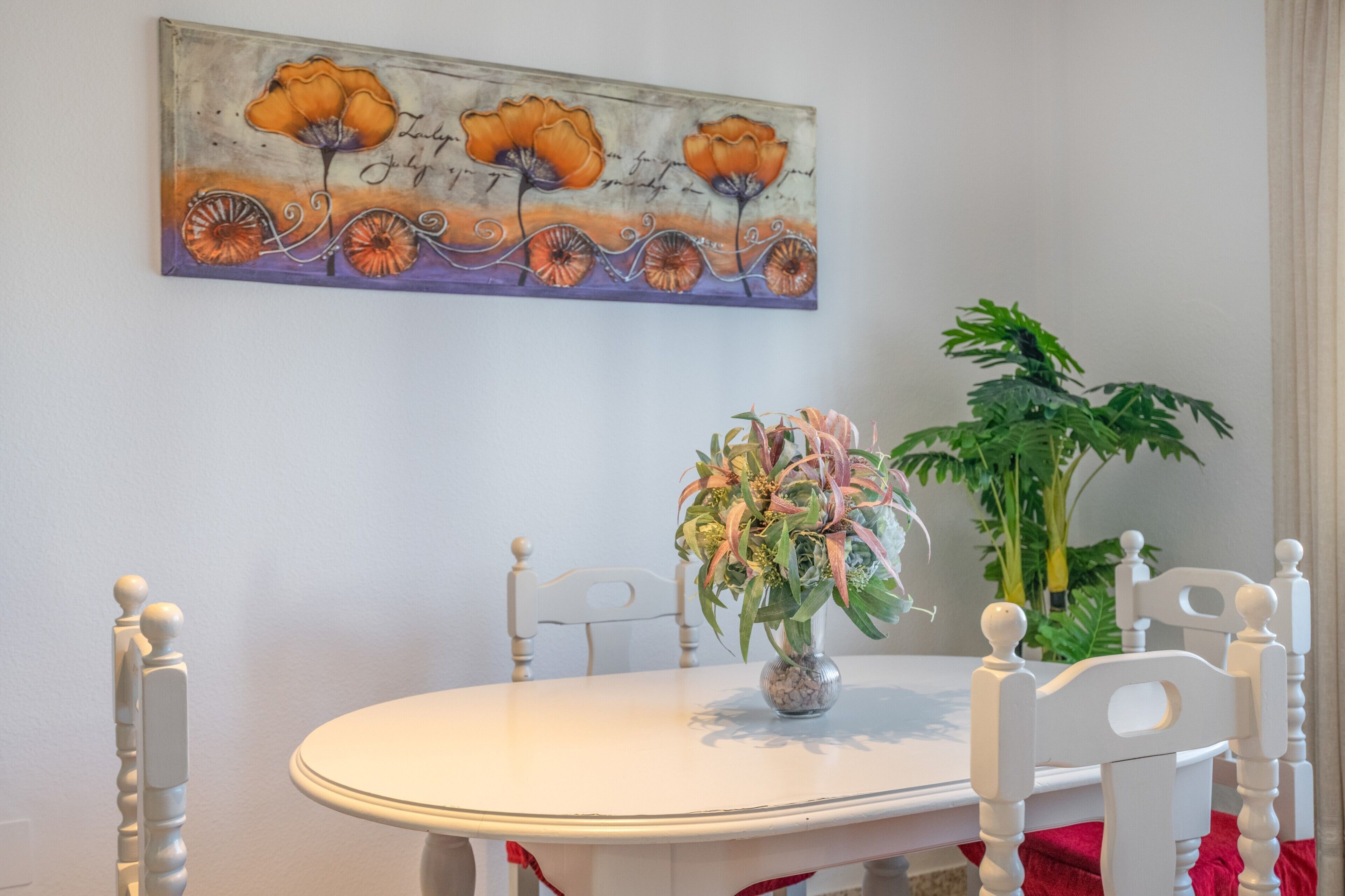 A dining table with chairs, a vase with flowers, a painting on the wall, and a potted plant.
