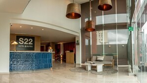 Un hall de hotel con mostrador de recepción, una zona de estar con sofá y mesa de centro, y grandes lámparas colgantes.