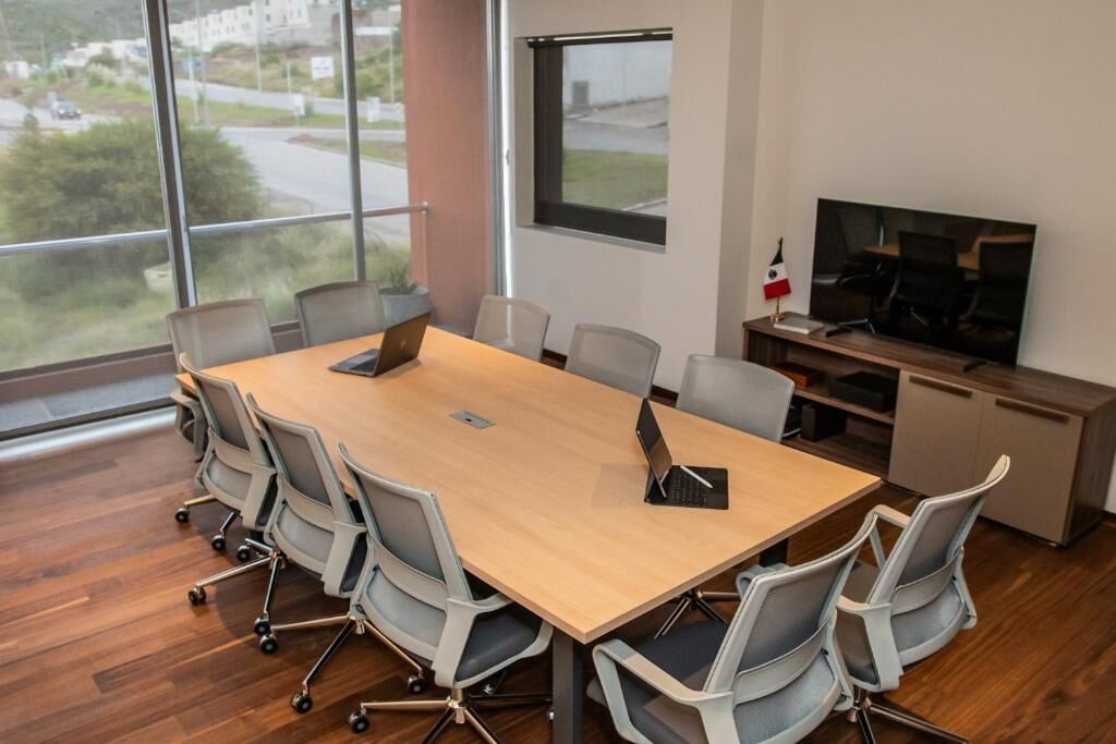 Un salón de conferencias con una mesa grande, seis sillas, un televisor y una ventana que ofrece vistas a una carretera y zonas verdes.