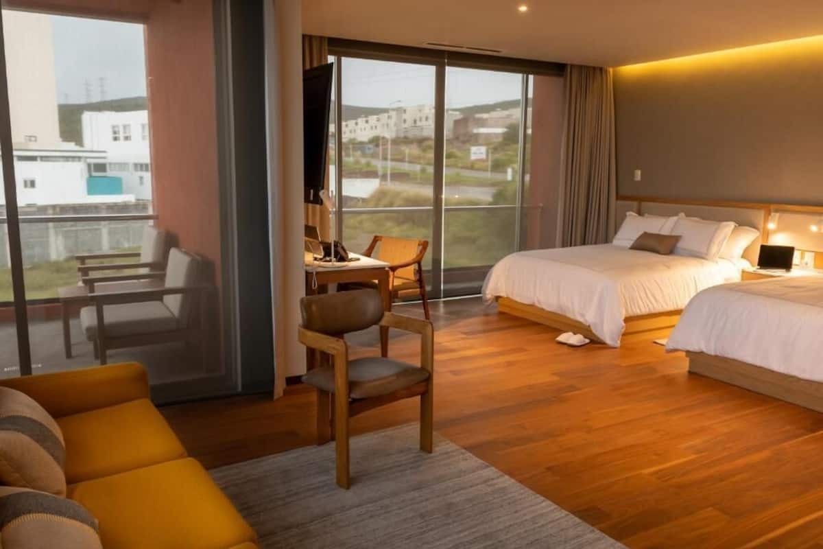 Une chambre d’hôtel avec un grand lit, un bureau avec un ordinateur portable, une chaise et un canapé. Il y a une grande fenêtre donnant sur des immeubles et un balcon.
