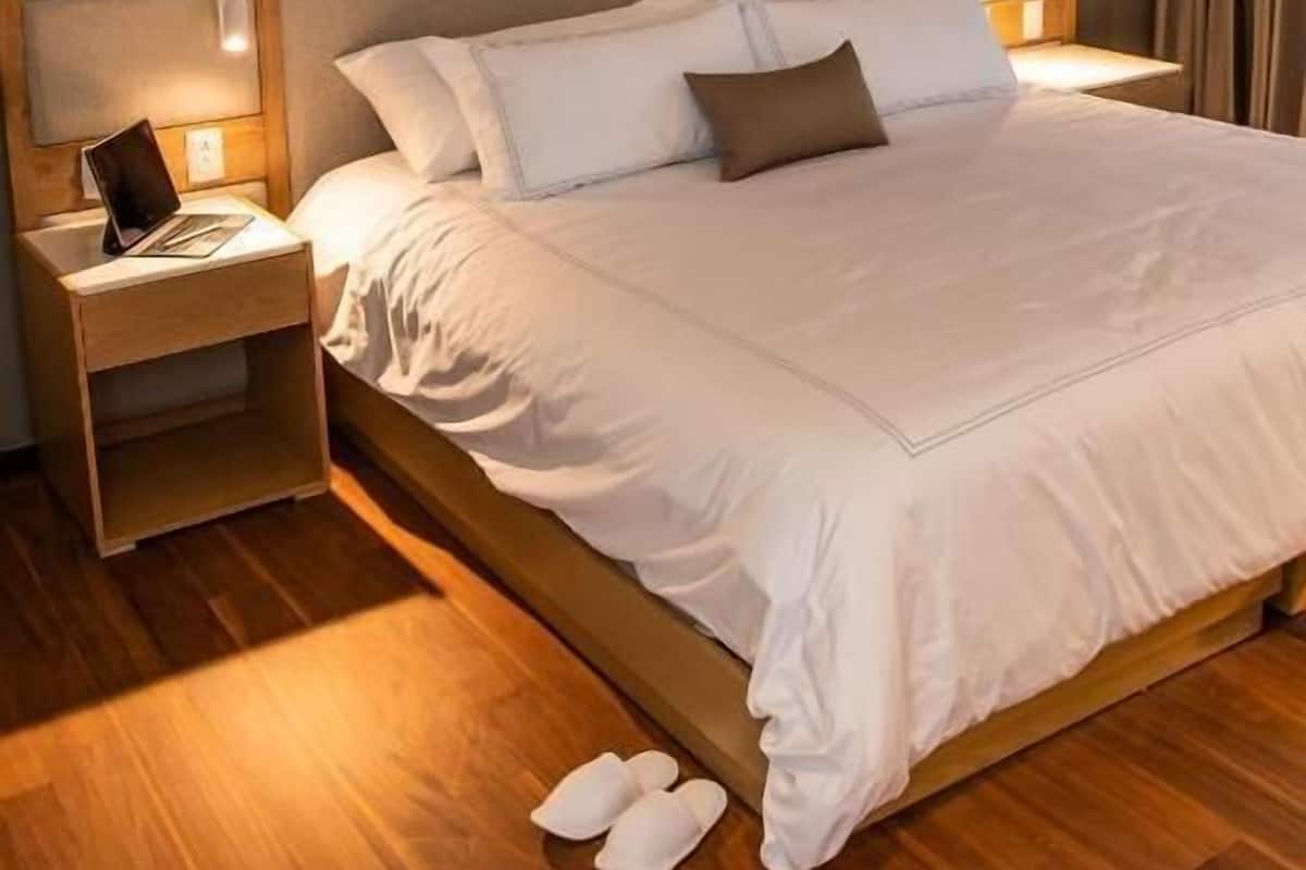 Un lit bien fait, avec du linge de lit blanc et une tête de lit en bois, une table de chevet avec une lampe, et une paire de pantoufles posées sur le sol.