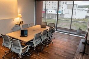 Un salón de conferencias con una gran mesa, seis sillas, una computadora portátil, una lámpara y vistas a edificios a través de amplias ventanas.