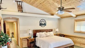 Een gezellige slaapkamer met een houten plafond, een bed met wit beddengoed, twee nachtkastjes met lampen, een houten bankje en een logo dat aan de muur is bevestigd.