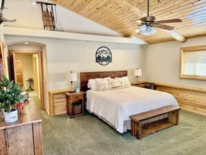 Ein gemütliches Schlafzimmer mit Holzbalkendecke, einem Bett mit weißer Bettwäsche, zwei Nachttischen mit Lampen, einer Holzbank und einem an der Wand befestigten Logo.