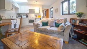 A cozy living room with a sofa, a wooden coffee table, and a dining table. There is a kitchen area with appliances and cabinets. - Goose Shed | Rural Farm Cottage | Cosy Fire (Stelling Minnis)