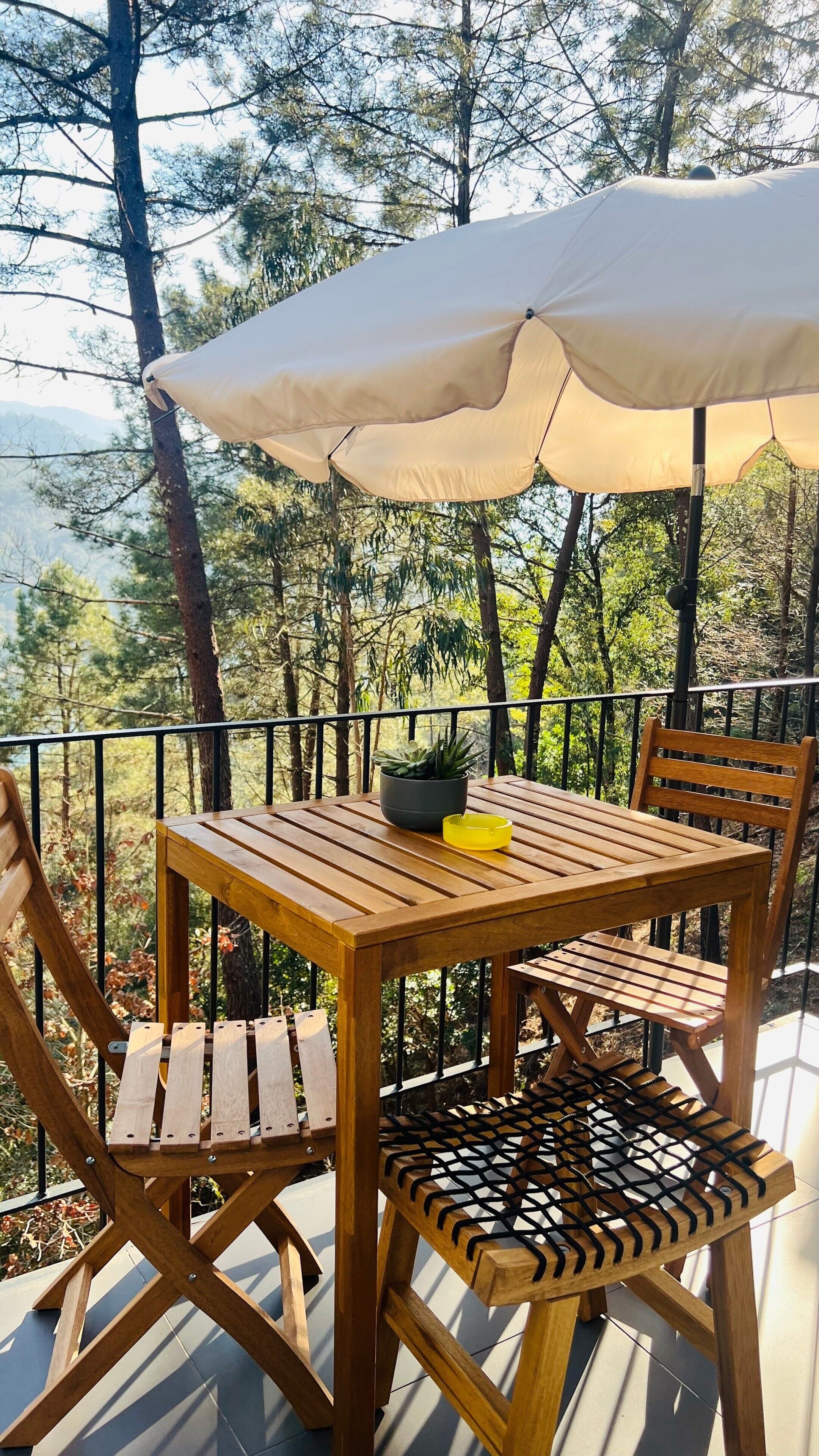 Mesa de madera con sillas y un paraguas grande en una terraza al aire libre, con vistas a árboles y montañas.