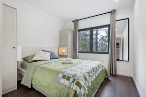 Ein Schlafzimmer mit Bett, Nachttisch, Fenster mit Vorhängen und Blick auf Bäume.