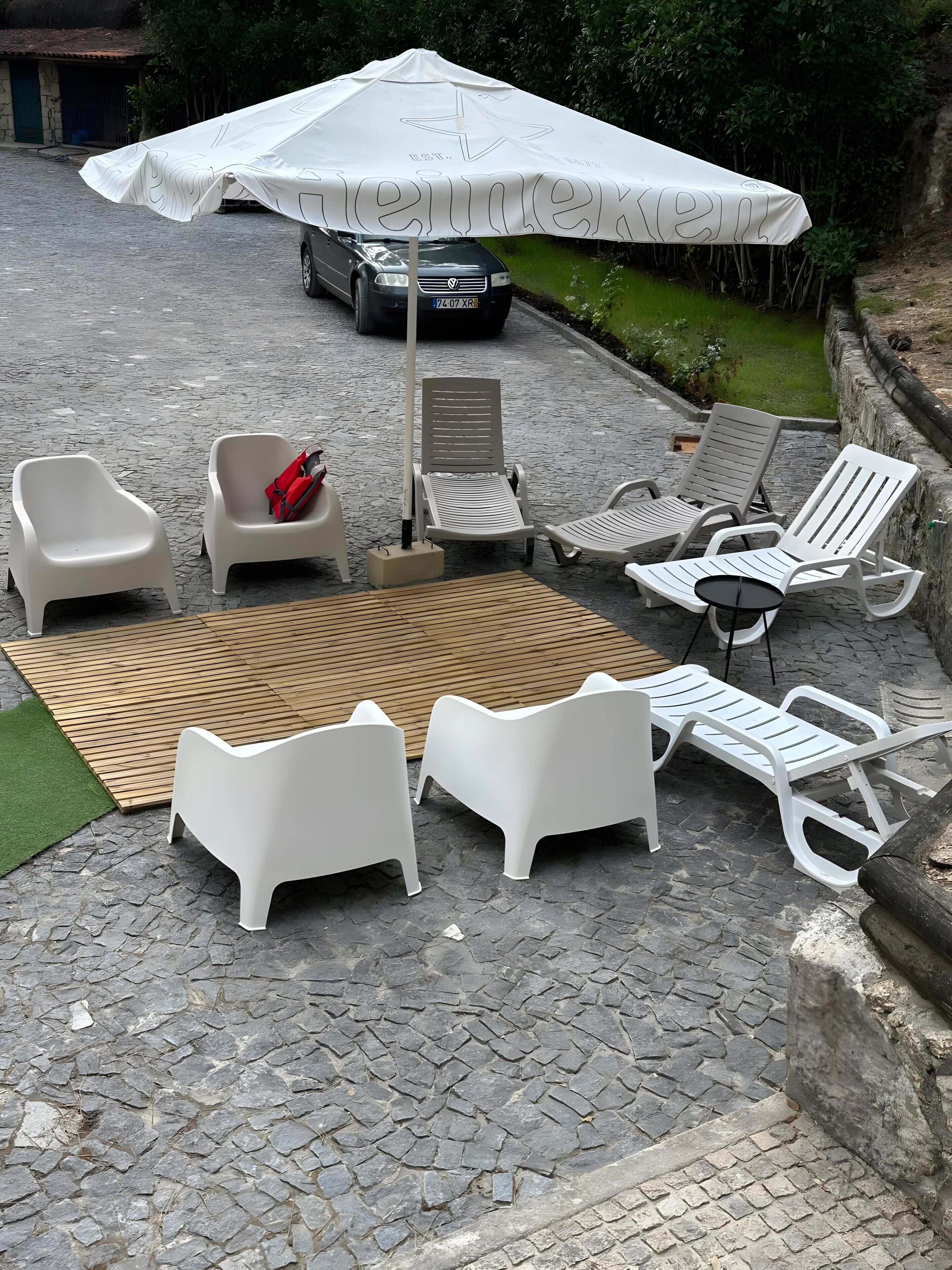 Eine Terrassenfläche mit Gartenmöbeln, darunter weiße Lounge-Stühle und ein Tisch, unter einem großen weißen Sonnenschirm.