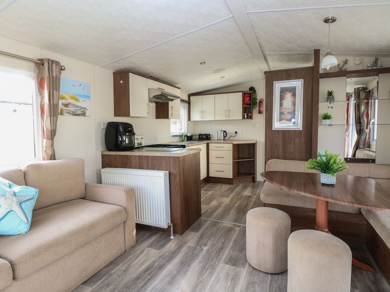 Eine kompakte Küchenzeile in einem Wohnmobil mit einer zentralen Kochinsel, einem Essbereich mit Holztisch und einem Wohnbereich mit Sofa und Hocker.