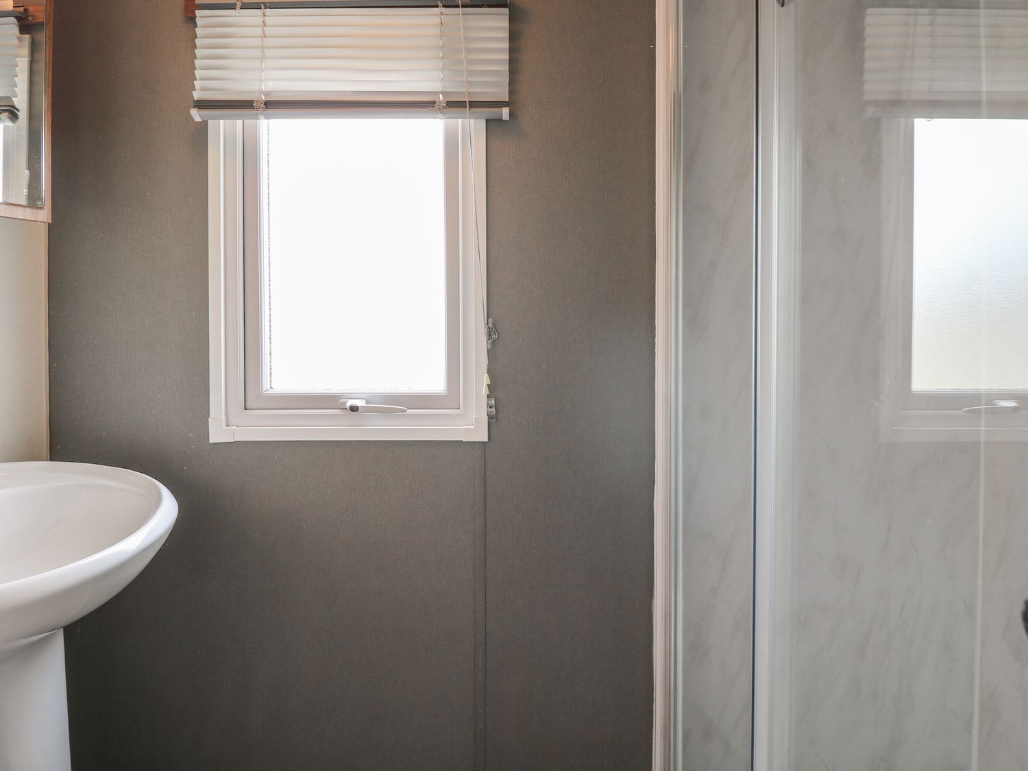 A bathroom with a white sink, a window with blinds, and a glass shower enclosure.