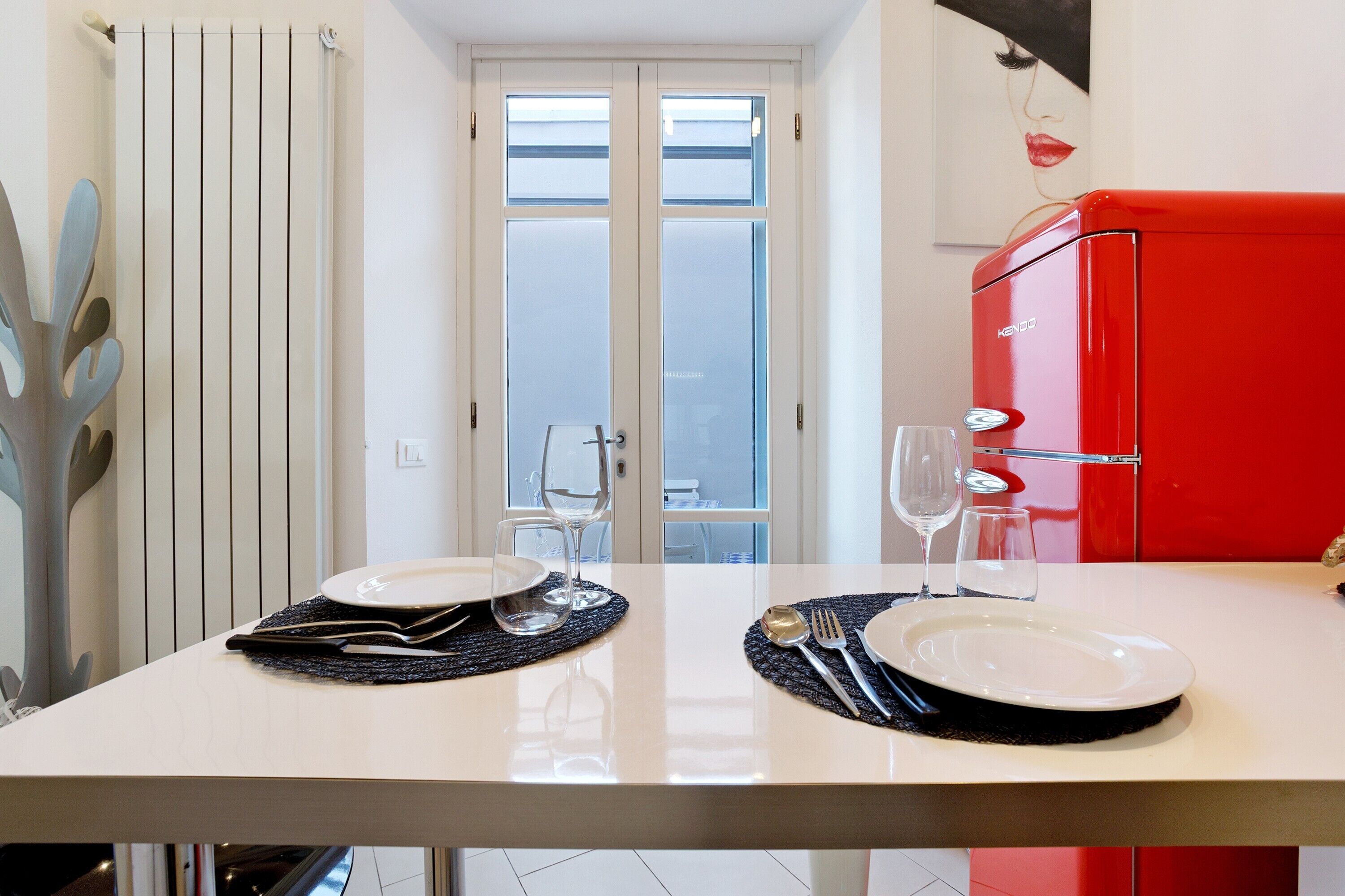 Una mesa de comedor dispuesta con platos, cubiertos y vasos, un electrodoméstico rojo, y vistas a una puerta con paneles de vidrio esmerilado.