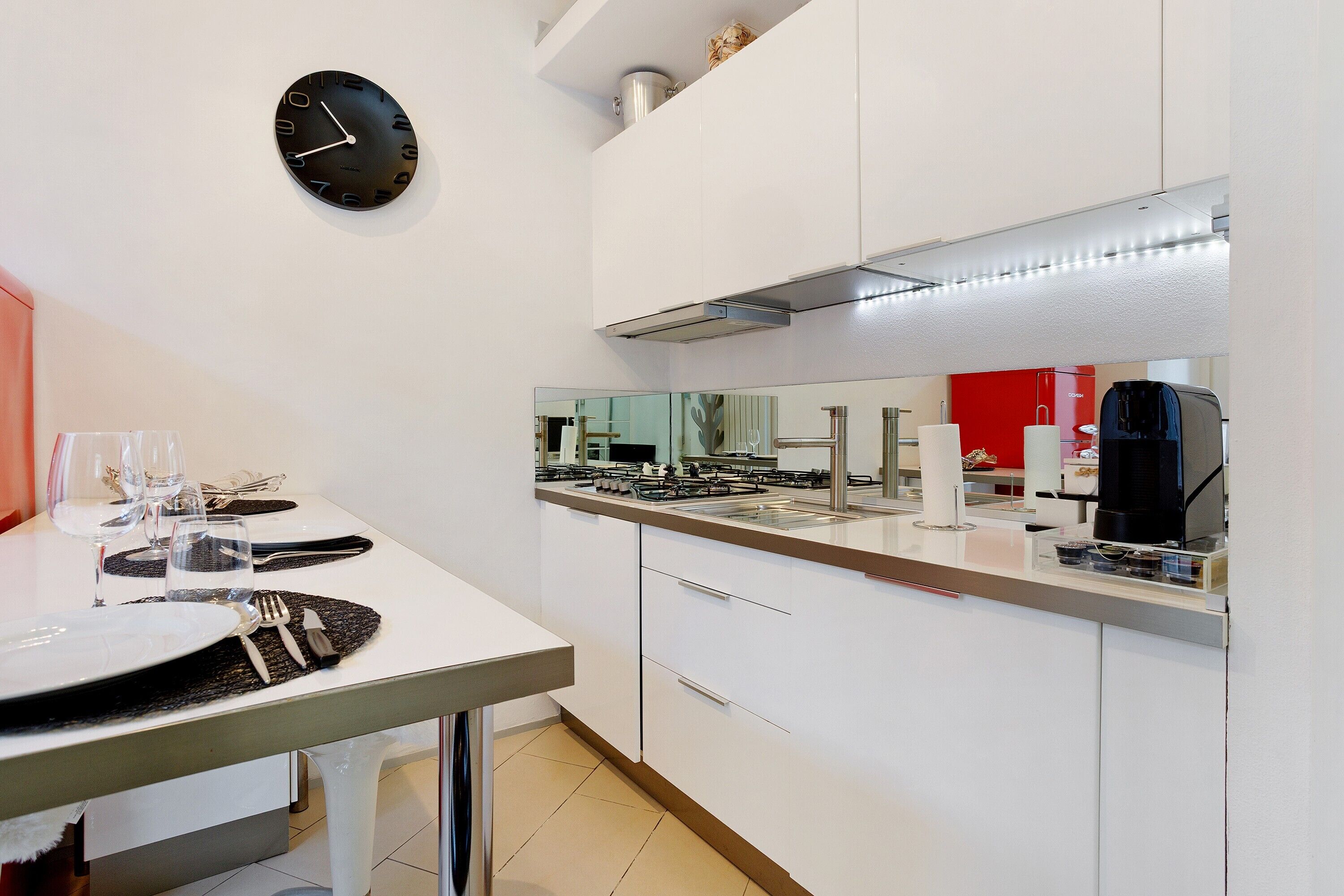A modern kitchen with white cabinets, a black clock, a stainless steel range hood, and a large mirror above the counter.