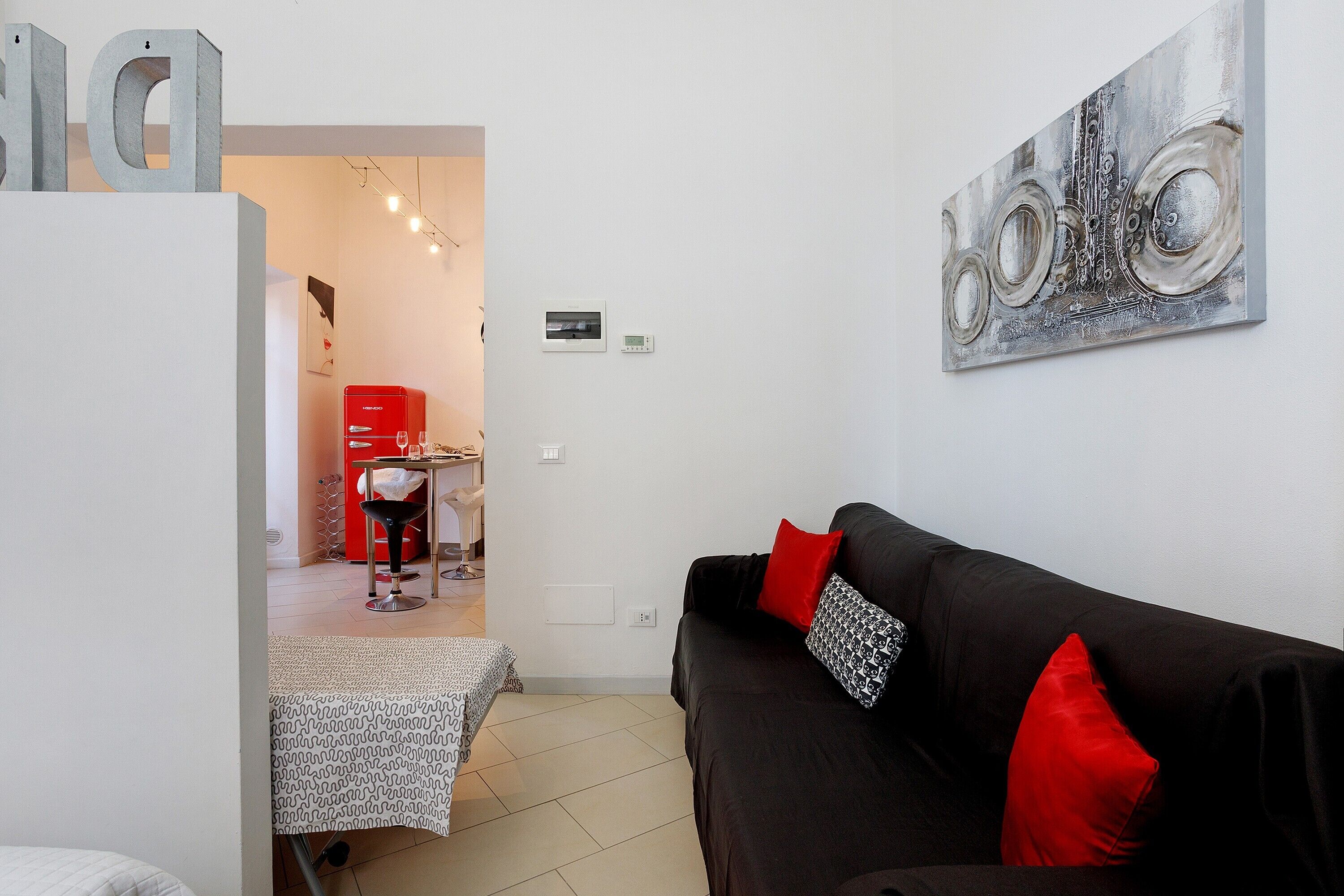 A modern living room with a black sofa, red and patterned pillows, a dining area with a red chair, and a wall-mounted abstract art piece.