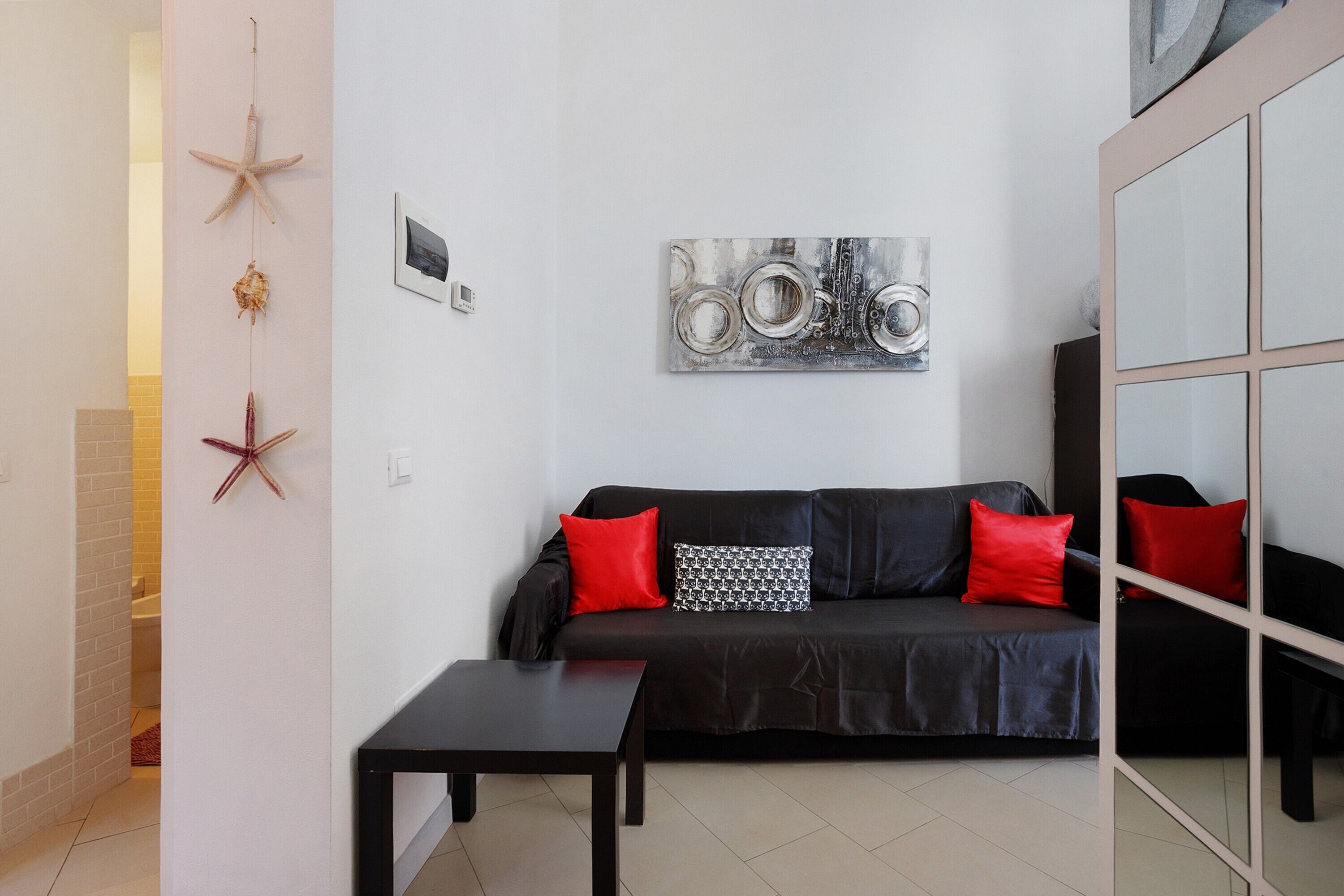 Een moderne woonkamer met een zwarte leren bank, rode en gemêleerde kussens, een salontafel en wanddecoratie, waaronder een zeester en een abstract ontwerp.