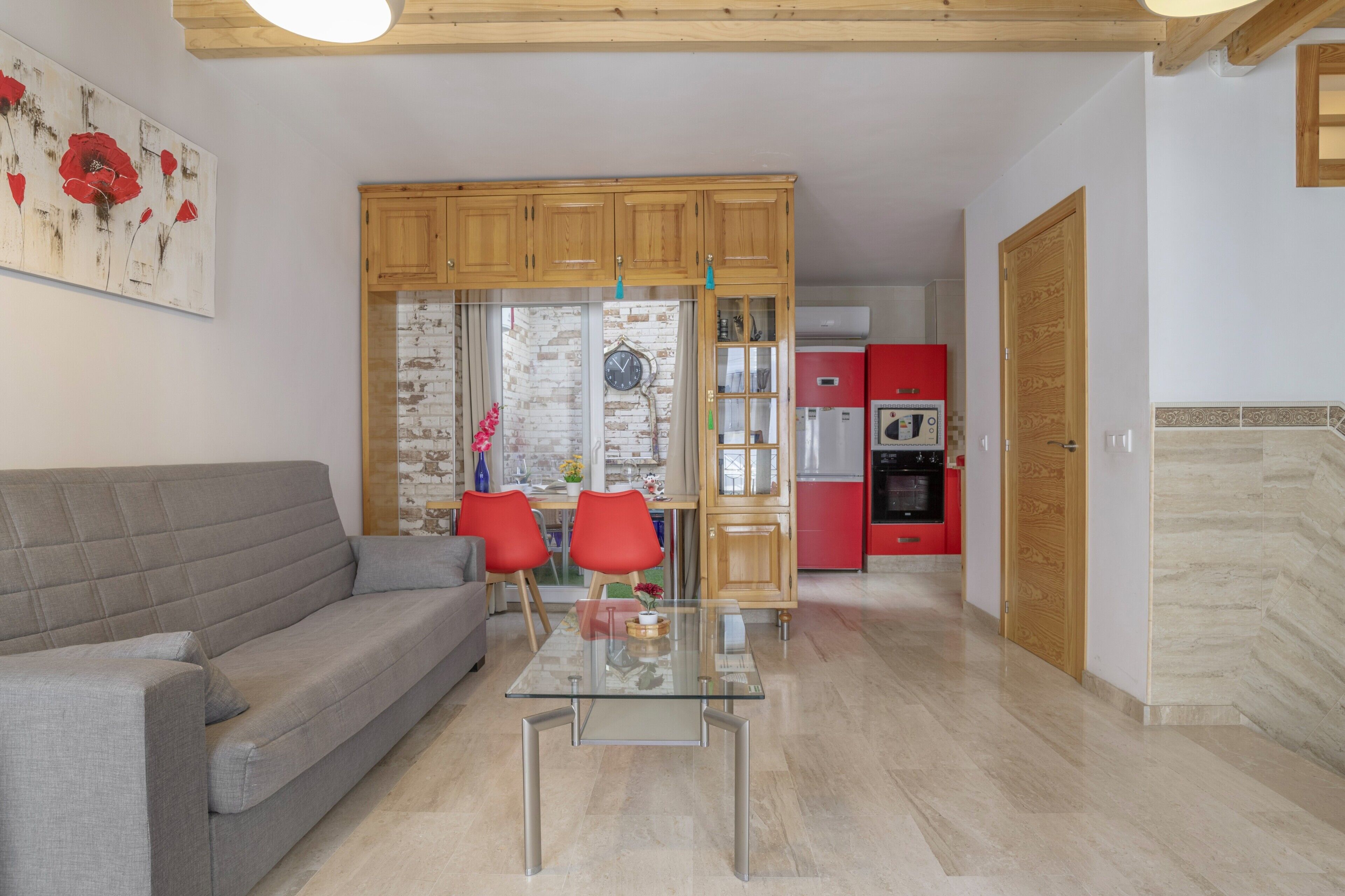 A living room with a sofa, a glass coffee table, and red chairs. There is a kitchen area with wooden cabinets and a red refrigerator.