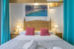 Un lit bien fait, avec deux oreillers, un tableau représentant un paysage côtier et deux lampes de chevet.