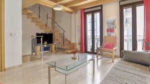 A spacious living area with a glass coffee table, a sofa, a striped armchair, a television, and a staircase.