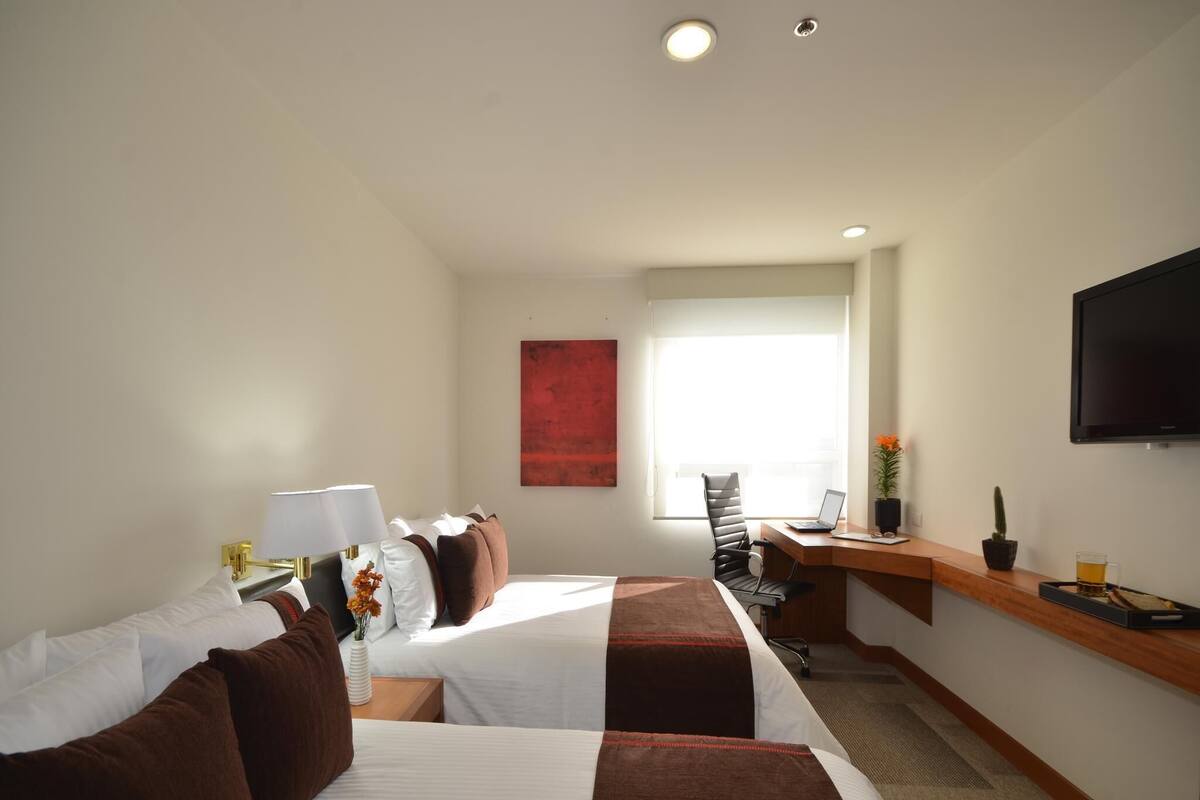 A hotel room with two beds, a desk with a laptop, a television, and a window with a view.