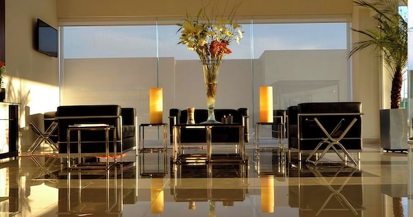 A modern lobby with a reflective floor, black leather seating, a tall vase with flowers, and a large window.