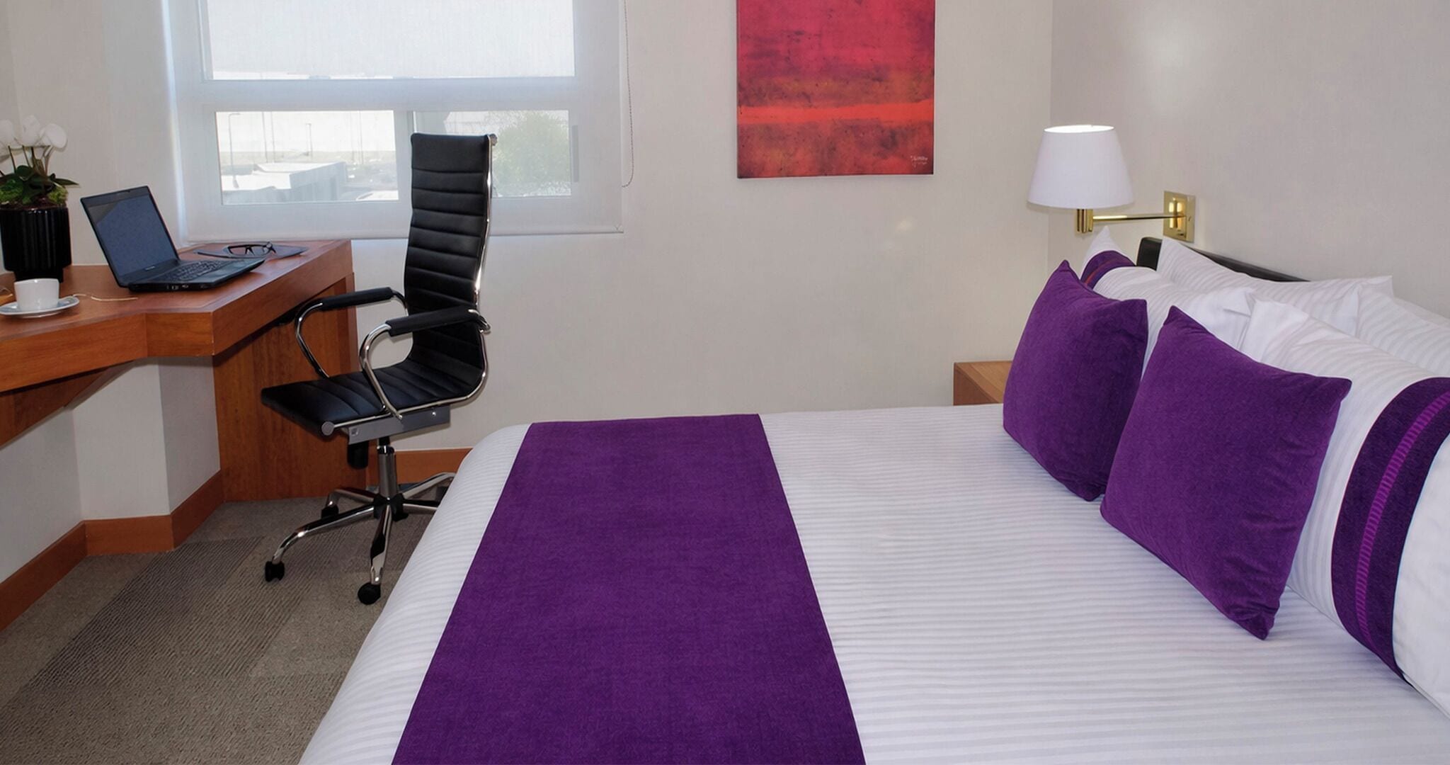 A hotel room with a bed, a desk with a laptop, a chair, a nightstand with a lamp, and a window.