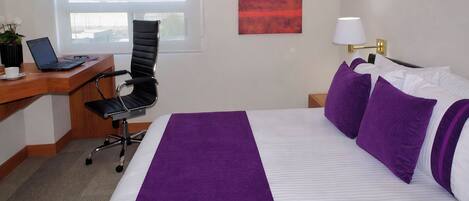 A hotel room with a bed, a desk with a laptop, a chair, a nightstand with a lamp, and a window.