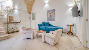 Un saló amb un sofà blau, dos butaques, una taula de centre i una televisió penjada a la paret, i una escala amb baranes de vidre.
