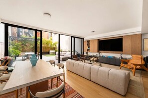 En moderne opholdsstue med store skydedøre til en terrasse, en sofa, et spisebord og et køkken med et fjernsyn.