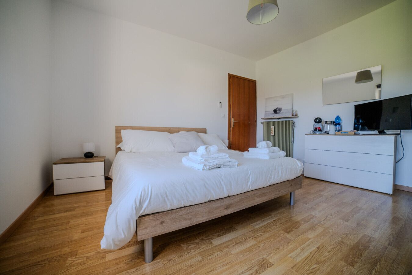 Una cama bien hecha con ropa de cama y toallas blancas, un cabecero de madera, una mesita de noche con lámpara, un aparador con espejo y un televisor sobre un mueble.