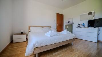 Una cama bien hecha con ropa de cama y toallas blancas, un cabecero de madera, una mesita de noche con lámpara, un aparador con espejo y un televisor sobre un mueble.