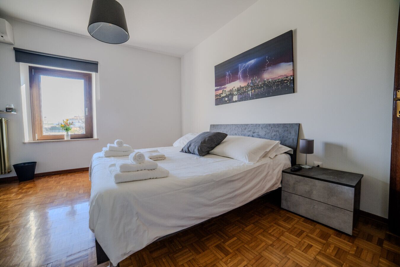 A modern bedroom with a bed, bedside table, and a cityscape painting.