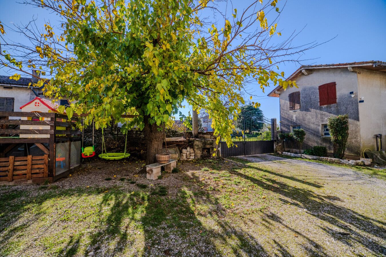 Une cour bordée d’arbres, avec une balançoire, un banc en pierre et une petite maison de jeu rustique.