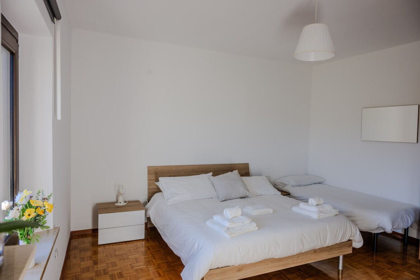 Une chambre à coucher avec un lit en bois, du linge de lit blanc, une table de chevet avec une lampe et une petite table de nuit ornée d’un vase de fleurs.