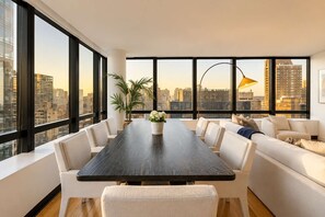 A modern dining room with a long table, white sofas, and a city view through large windows. - Skyline V | 26th-floor 3BR w/Doorman, Gym, and Pool (New York)